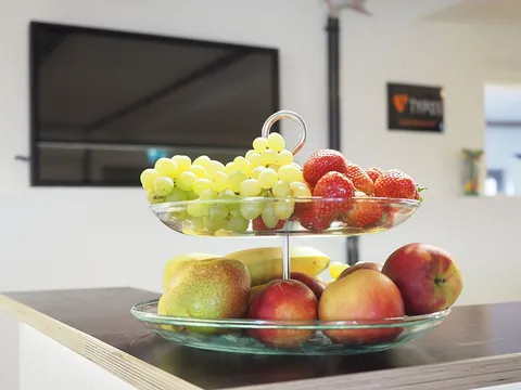 Obstschale mit Trauben, Äpfeln und Bananen auf einem Tisch im Büro von e-pixler, im Hintergrund ein Fernseher.