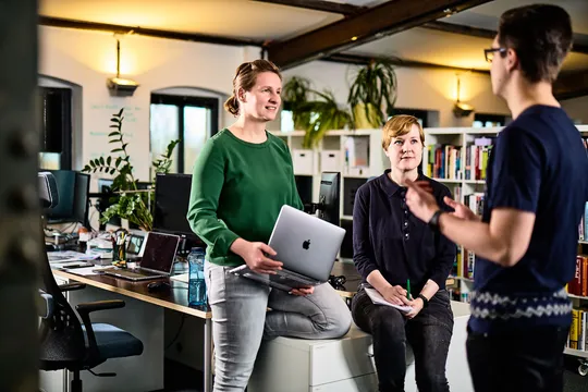 Zwei e-pixler-Mitarbeiterinnen im Gespräch, eine hält einen Laptop, während ein Kollege spricht.