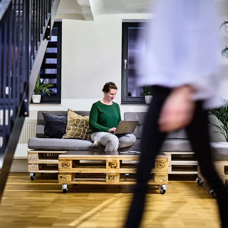 Mitarbeiterin bei e-pixler arbeitet mit einem Laptop auf einer Couch aus Paletten in einem modernen Büro mit Pflanzen und Holzfußboden.