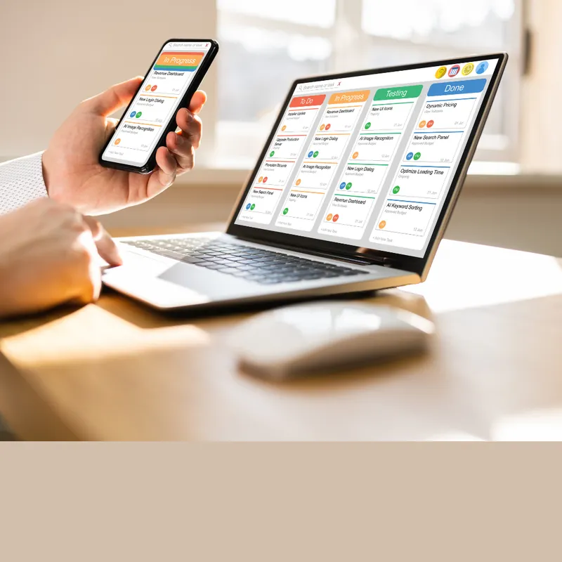 Person arbeitet mit einem Smartphone und einem Laptop, die beide eine digitale Organisationssoftware mit farblich gekennzeichneten Aufgaben und Kalenderansichten anzeigen.